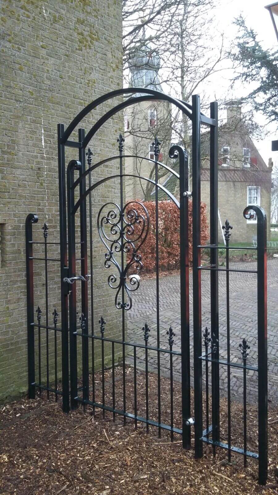 Impressie van een rozenboog met hekje aan beide zijden. De rozenboog is zwart, van staal en heeft sierlijke krullen en ornamenten.