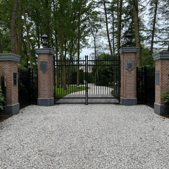 Een statige, zwarte metalen toegangspoort tussen vier gemetselde bakstenen pilaren. De middelste twee pilaren zijn voorzien van een lantaarnlamp aan de bovenkant en een grijs wapenschild aan de voorzijde.